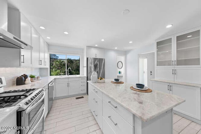 kitchen featuring light stone counters, stainless steel appliances, wall chimney range hood, white cabinets, and a center island