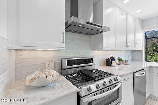 kitchen featuring light stone countertops, wall chimney exhaust hood, tasteful backsplash, white cabinets, and appliances with stainless steel finishes