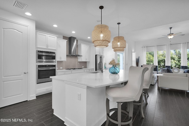 kitchen featuring white cabinetry, an island with sink, sink, hanging light fixtures, and wall chimney exhaust hood