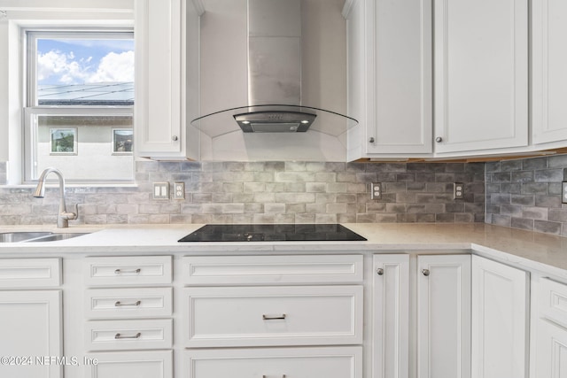 kitchen featuring wall chimney range hood, backsplash, white cabinetry, black electric stovetop, and sink