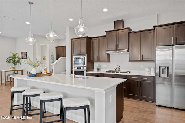 kitchen featuring hanging light fixtures, appliances with stainless steel finishes, light wood-type flooring, and a kitchen island with sink