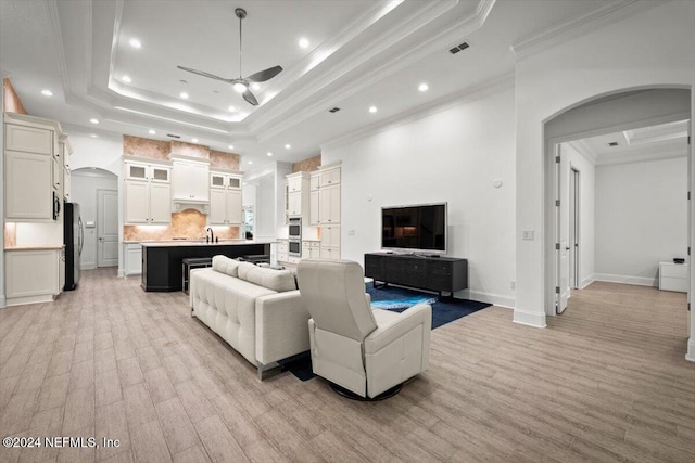 living room with ceiling fan, a raised ceiling, light wood-type flooring, ornamental molding, and sink