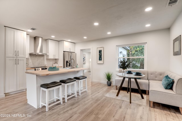 kitchen featuring wall chimney range hood, a kitchen breakfast bar, light hardwood / wood-style flooring, stainless steel fridge, and white cabinets