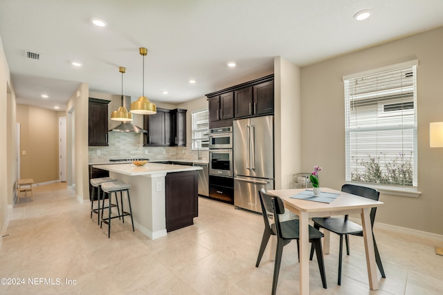 kitchen with a kitchen island, wall chimney range hood, hanging light fixtures, stainless steel appliances, and backsplash