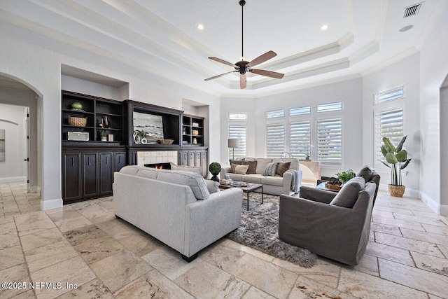 living room featuring a raised ceiling, crown molding, ceiling fan, built in shelves, and a tiled fireplace