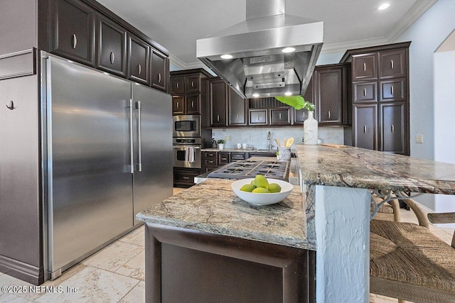 kitchen featuring built in appliances, light stone countertops, tasteful backsplash, a kitchen bar, and island exhaust hood