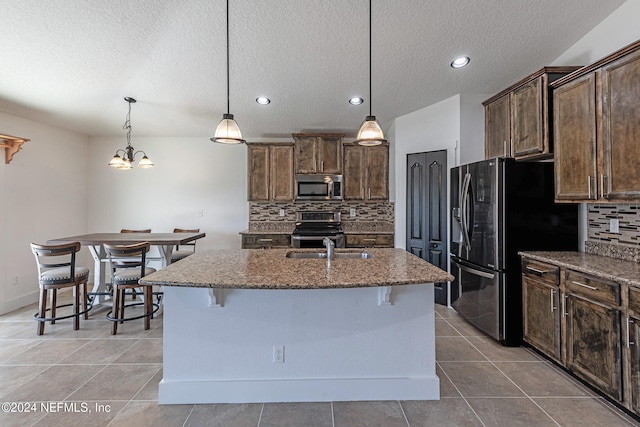 kitchen featuring a breakfast bar, appliances with stainless steel finishes, a center island with sink, decorative light fixtures, and dark stone counters