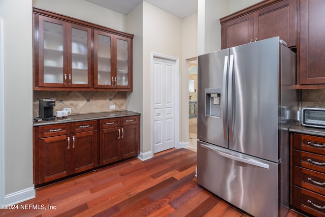 kitchen featuring stainless steel fridge, dark wood-style floors, glass insert cabinets, dark stone countertops, and backsplash