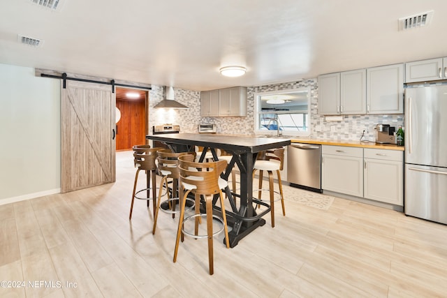 kitchen featuring light wood-type flooring, a barn door, a kitchen bar, stainless steel appliances, and wall chimney exhaust hood