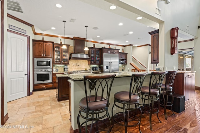 kitchen with a large island with sink, a kitchen breakfast bar, built in appliances, light stone countertops, and ornamental molding