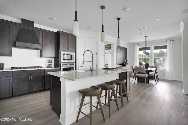 kitchen featuring a center island with sink, appliances with stainless steel finishes, wood-type flooring, and wall chimney exhaust hood