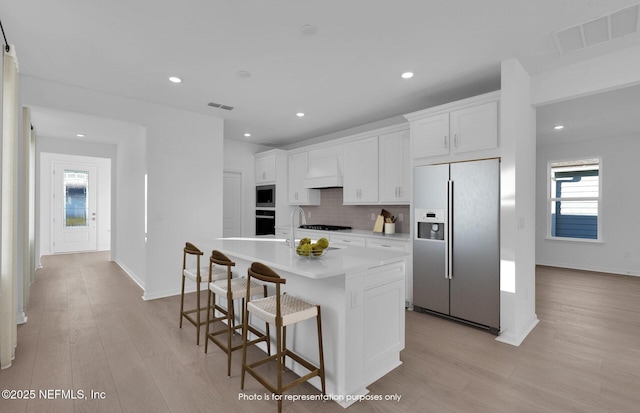 kitchen featuring visible vents, black appliances, an island with sink, decorative backsplash, and custom exhaust hood