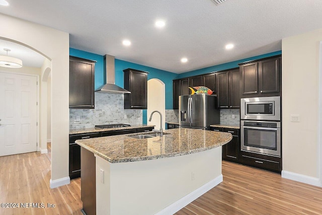 kitchen featuring wall chimney range hood, a center island with sink, appliances with stainless steel finishes, light hardwood / wood-style flooring, and sink