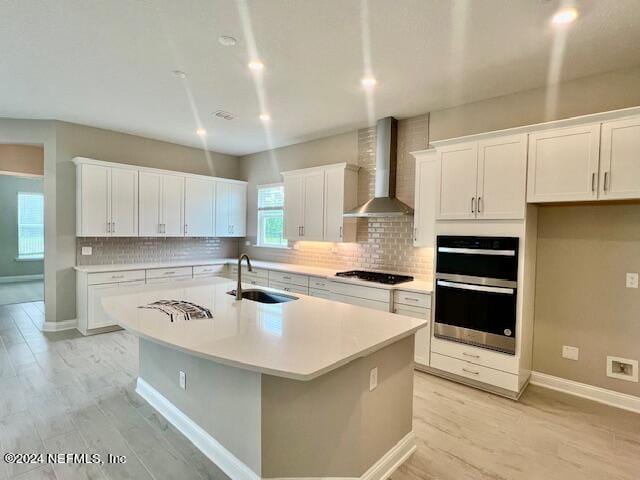 kitchen with sink, double oven, wall chimney exhaust hood, white cabinets, and a center island with sink