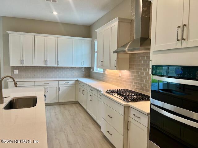 kitchen with decorative backsplash, white cabinetry, light hardwood / wood-style flooring, wall chimney exhaust hood, and stainless steel appliances