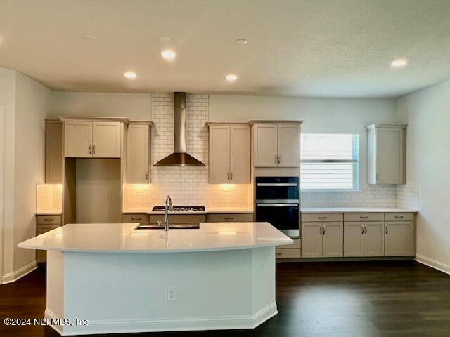 kitchen with dark hardwood / wood-style flooring, backsplash, an island with sink, wall chimney exhaust hood, and double oven