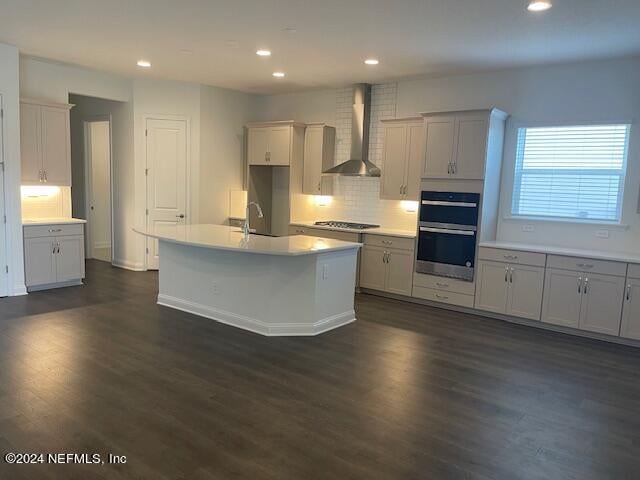 kitchen with double oven, dark hardwood / wood-style flooring, wall chimney exhaust hood, gas cooktop, and a kitchen island with sink