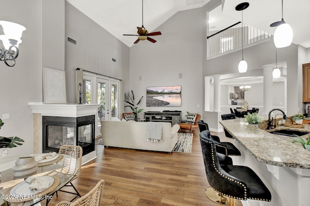 living room featuring sink, light wood-type flooring, ceiling fan with notable chandelier, high vaulted ceiling, and a multi sided fireplace