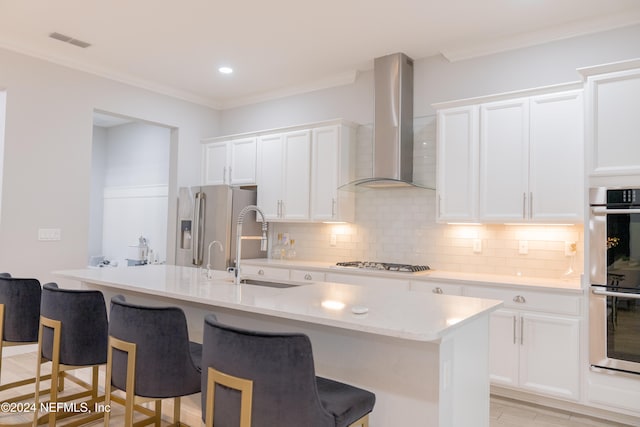 kitchen featuring appliances with stainless steel finishes, an island with sink, wall chimney range hood, and white cabinetry