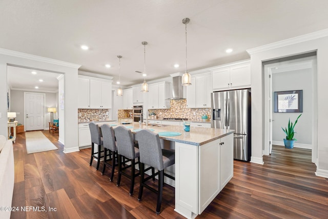 kitchen featuring white cabinets, stainless steel appliances, wall chimney exhaust hood, a center island with sink, and backsplash