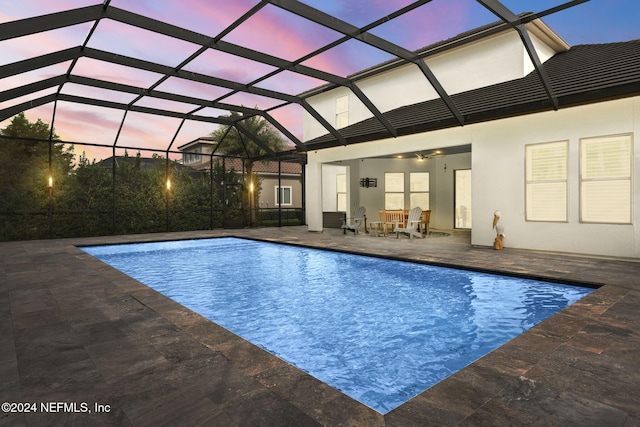 pool at dusk with glass enclosure, a patio, and ceiling fan