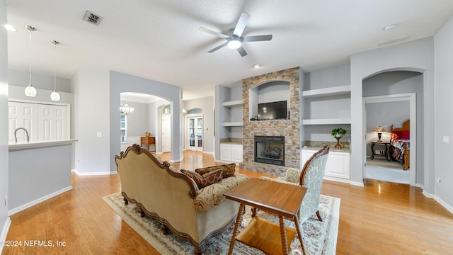 living room featuring a fireplace, light wood-type flooring, built in features, a textured ceiling, and ceiling fan with notable chandelier