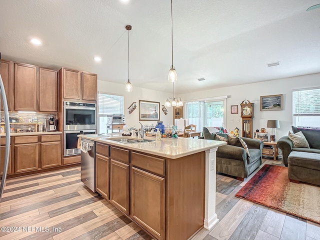 kitchen with appliances with stainless steel finishes, sink, light wood-type flooring, decorative light fixtures, and a kitchen island with sink