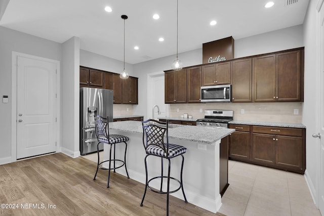 kitchen featuring hanging light fixtures, an island with sink, light stone countertops, light wood-type flooring, and stainless steel appliances