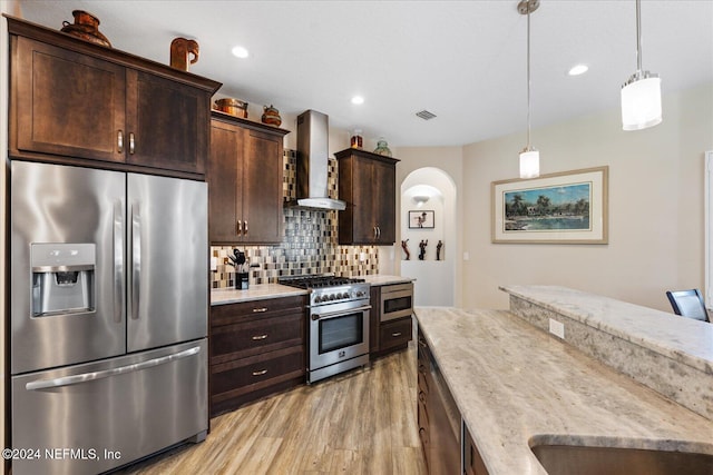 kitchen featuring wall chimney range hood, appliances with stainless steel finishes, light wood-type flooring, pendant lighting, and dark brown cabinetry