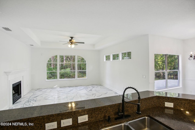 kitchen featuring a raised ceiling, ceiling fan, a high end fireplace, dark stone counters, and sink