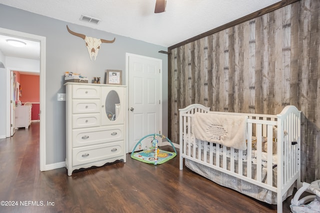 bedroom with a textured ceiling, a crib, dark wood-type flooring, and ceiling fan