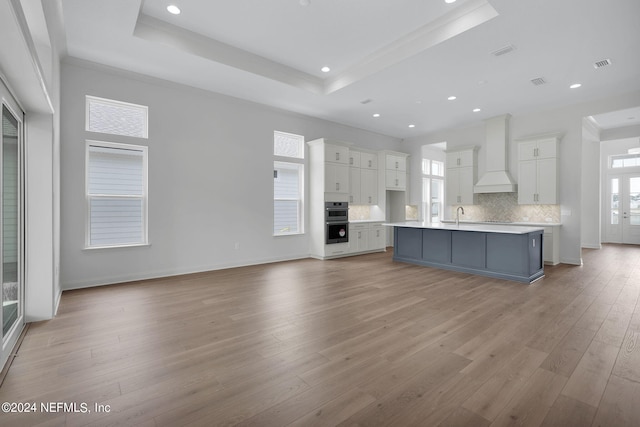 kitchen featuring light wood-type flooring, a tray ceiling, white cabinets, custom range hood, and a kitchen island with sink