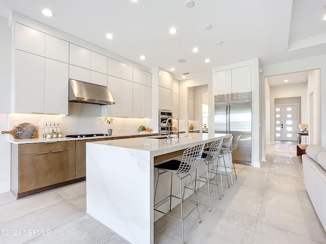 kitchen featuring white cabinetry, a kitchen island with sink, built in fridge, decorative backsplash, and exhaust hood