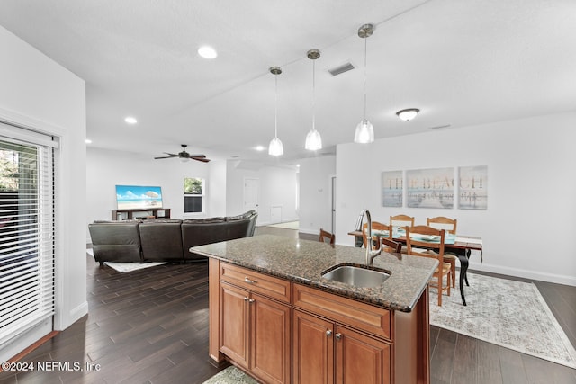 kitchen featuring a center island with sink, a sink, brown cabinetry, dark stone counters, and pendant lighting