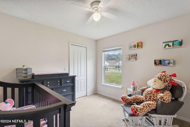 bedroom featuring ceiling fan, a textured ceiling, a nursery area, a closet, and light colored carpet