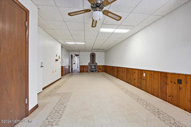 empty room with a wood stove, a drop ceiling, wooden walls, and ceiling fan