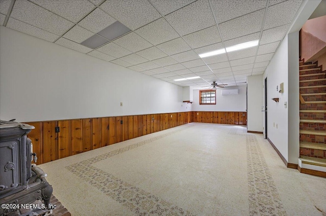 basement featuring wood walls, a drop ceiling, ceiling fan, and a wood stove