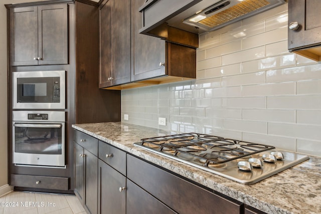 kitchen featuring stainless steel appliances, light stone counters, dark brown cabinetry, tasteful backsplash, and custom range hood