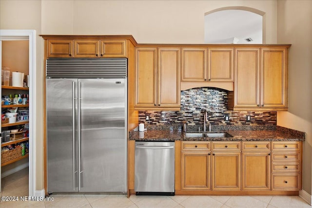 kitchen with appliances with stainless steel finishes, backsplash, sink, dark stone countertops, and light tile patterned flooring