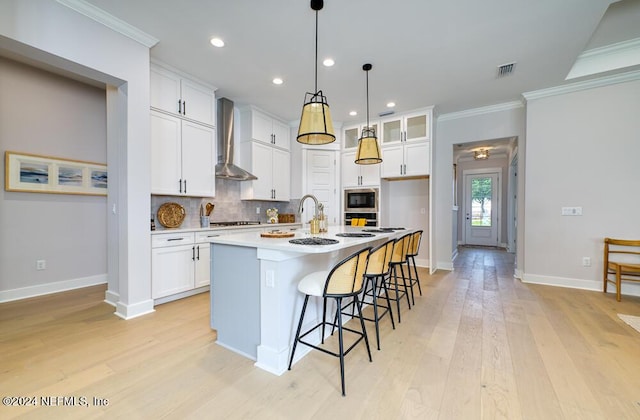 kitchen with an island with sink, white cabinetry, and wall chimney range hood