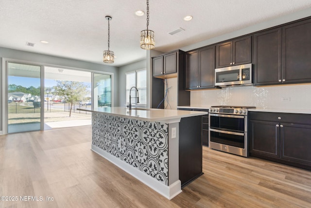 kitchen with tasteful backsplash, light hardwood / wood-style floors, decorative light fixtures, a kitchen island with sink, and appliances with stainless steel finishes