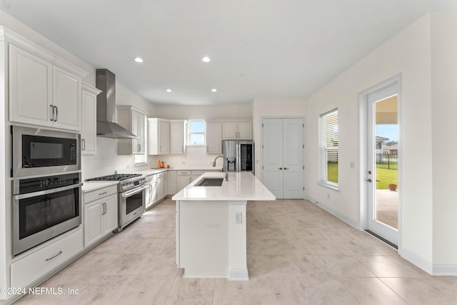 kitchen with appliances with stainless steel finishes, wall chimney exhaust hood, sink, a center island with sink, and white cabinets