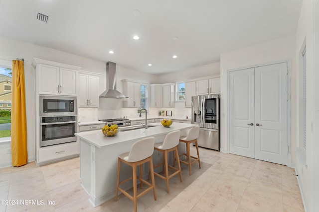 kitchen with appliances with stainless steel finishes, wall chimney exhaust hood, sink, white cabinets, and an island with sink