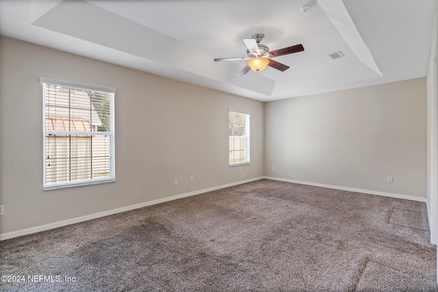 empty room with a wealth of natural light, ceiling fan, carpet flooring, and a raised ceiling