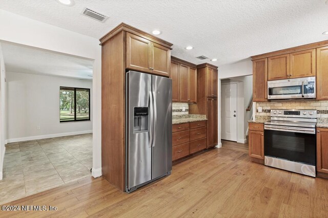 kitchen with light stone counters, a textured ceiling, tasteful backsplash, light wood-type flooring, and appliances with stainless steel finishes