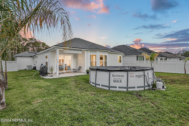 back house at dusk with a patio, a lawn, a covered pool, and cooling unit