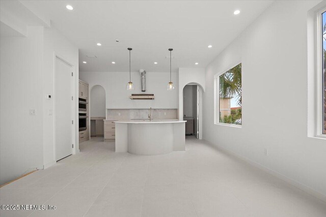 kitchen with light tile patterned floors, stainless steel double oven, an island with sink, hanging light fixtures, and sink