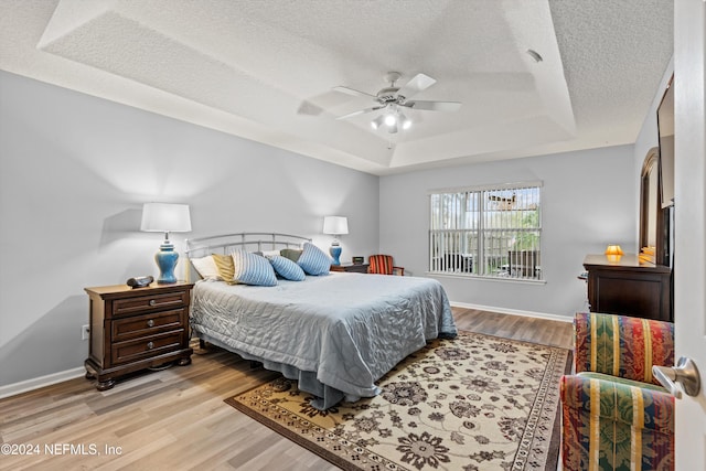 bedroom with a textured ceiling, light hardwood / wood-style floors, ceiling fan, and a raised ceiling