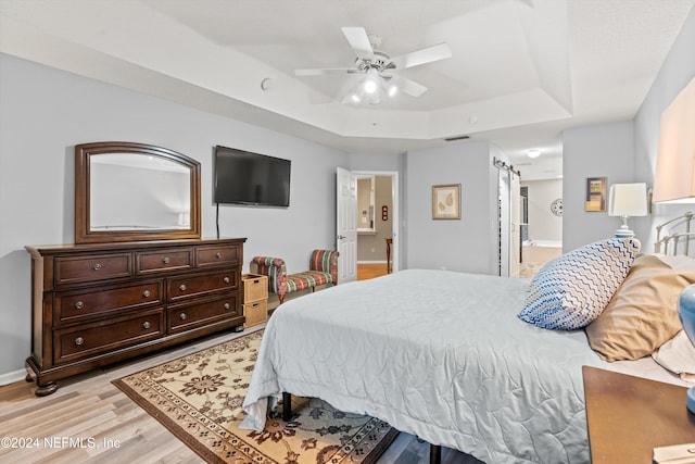 bedroom featuring ensuite bath, light wood-type flooring, a barn door, a tray ceiling, and ceiling fan