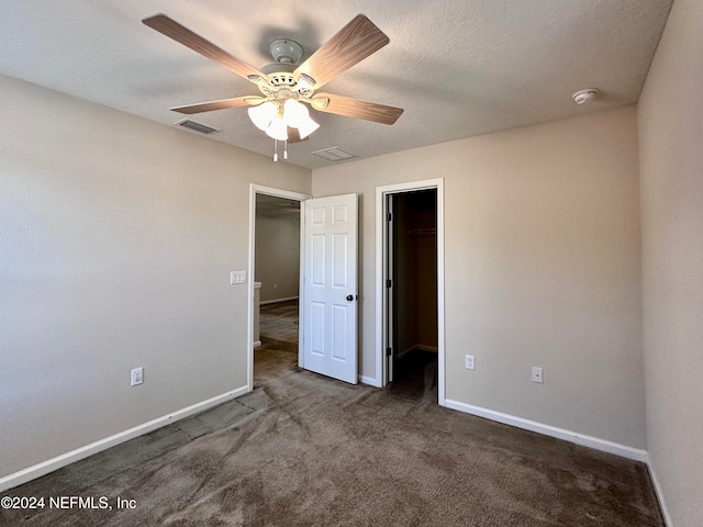 unfurnished bedroom featuring a spacious closet, a textured ceiling, dark carpet, a closet, and ceiling fan